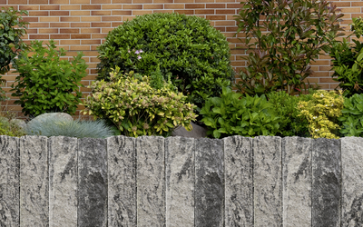Anthrazitfarbene Palisadenmauer aus Naturstein in einem Gartenbeet vor Hecke und Ziegelwand