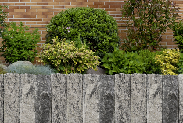 Anthrazitfarbene Palisadenmauer aus Naturstein in einem Gartenbeet vor Hecke und Ziegelwand