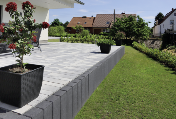 anthrazitfarbene Palisaden als Terrassenstützmauer neben gepflasterter Terrasse im Garten