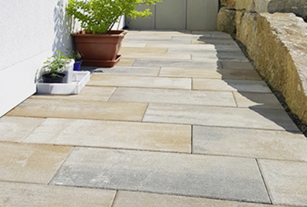 Terrassenplatten in Beige-Grau entlang eines Hauswegs, mit Pflanzkübeln und Natursteinmauer