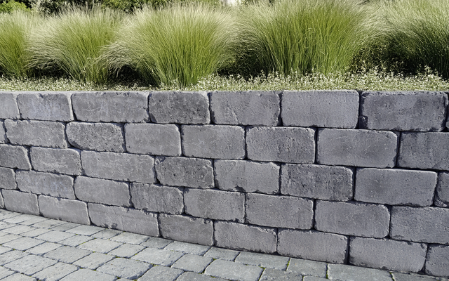 Graue Mauer mit Pflasterfläche davor und Ziergräsern dahinter