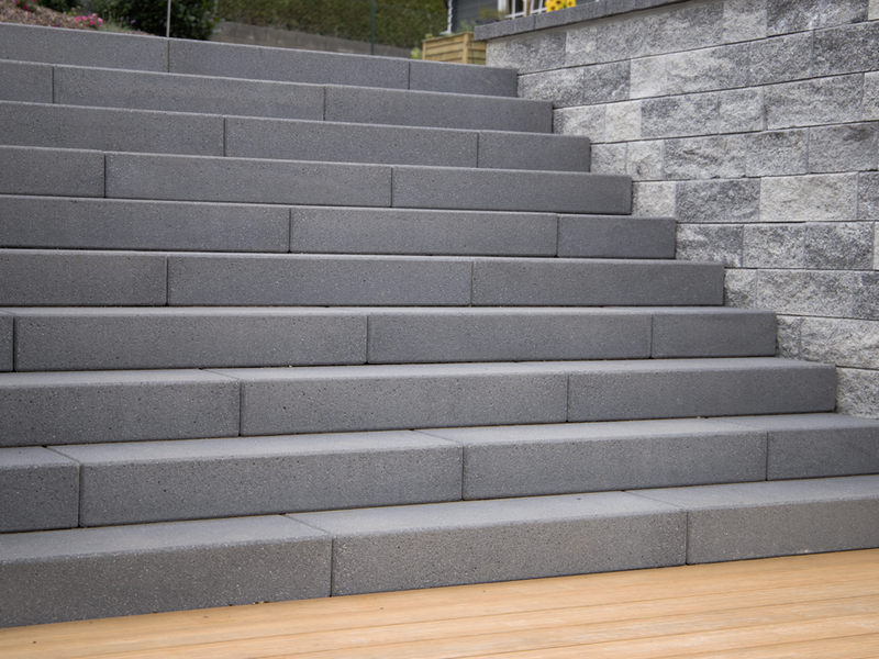Anthrazitfarbenen Blockstufen als Treppe im Außenbereich, vor einer Natursteinmauer und Holzterrasse