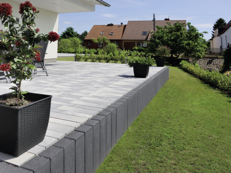 anthrazitfarbene Palisaden als Terrassenstützmauer neben gepflasterter Terrasse im Garten