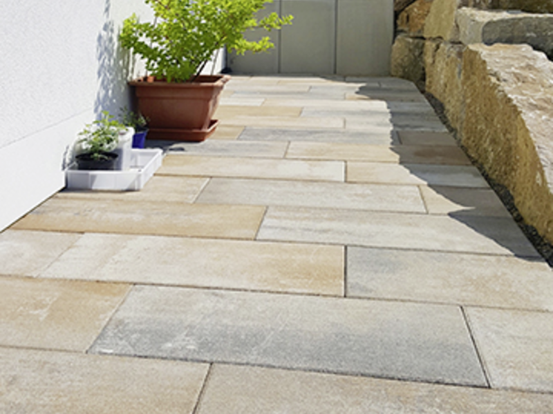 Terrassenplatten in Beige-Grau entlang eines Hauswegs, mit Pflanzkübeln und beiger Mauer