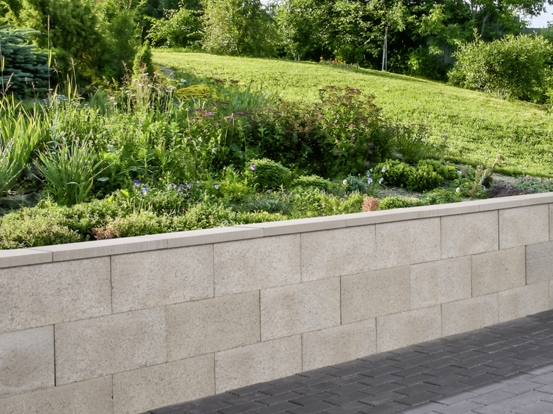 Grau gemauerte Mauer mit Abdeckplatte vor bepflanzter Gartenanlage und gepflastertem Weg