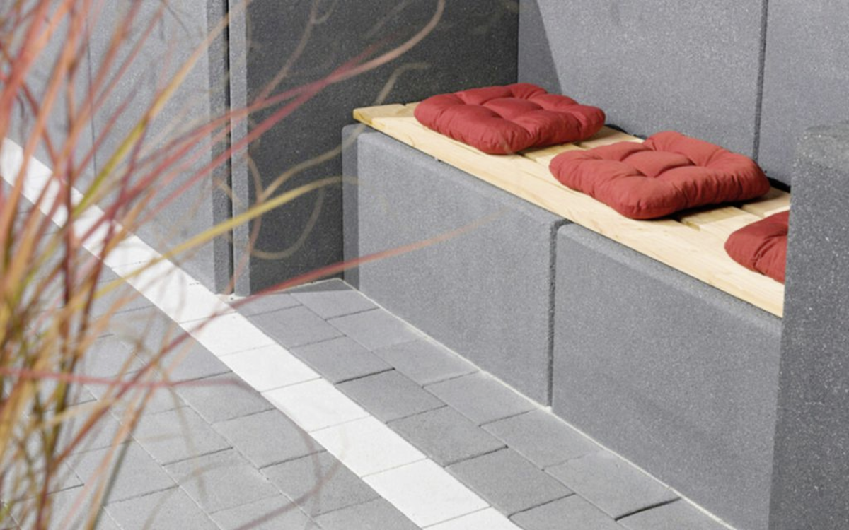 Grauer Stützwinkel mit Holzsitzbank und roten Sitzkissen an einer gepflasterten Gartenfläche