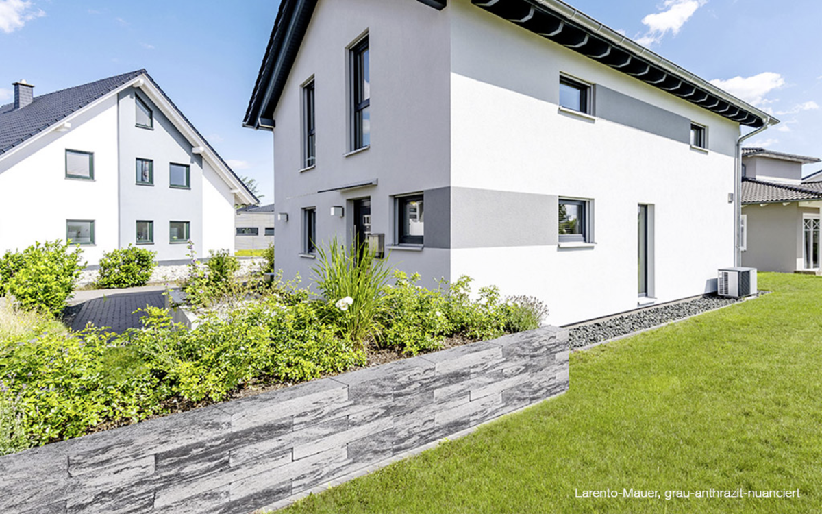 Graue Steinmauer als Beetbegrenzung vor einem gepflegten Garten mit Rasen und Wohnhaus